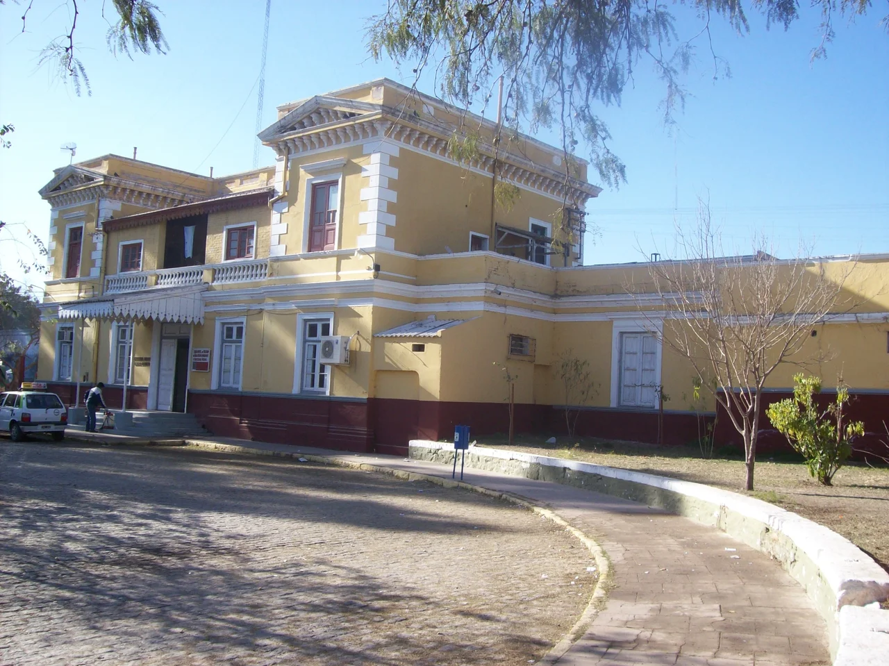 Frente de la antigua estación Santiago del Estero, con las vías levantadas. A la izquierda de la puerta principal se encuentra la leyenda "Ferrocarril Nacional General Belgrano" y a la derecha un cartel que indica "Subdirección de calidad de vida, medio ambiente y contralor municipal".