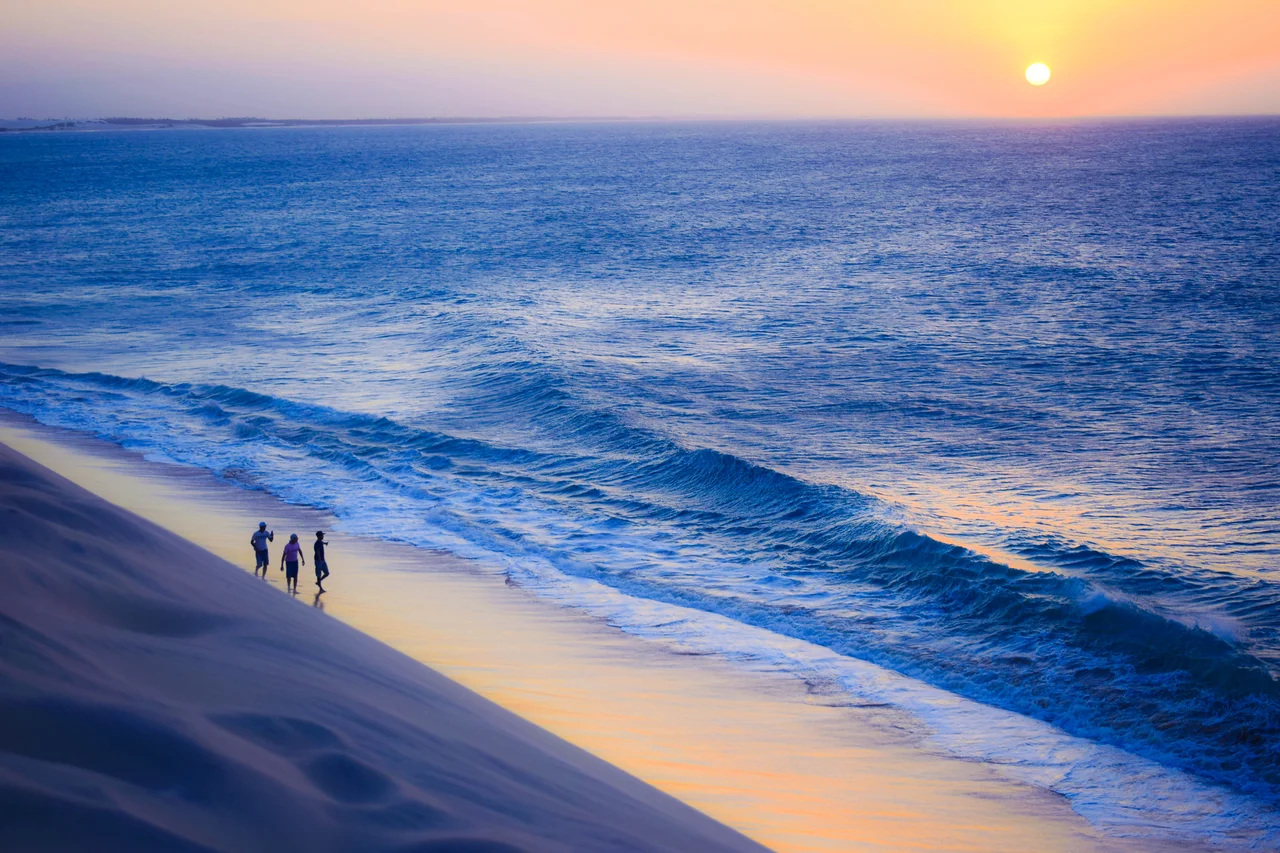 A vista do pôr do sol da maior duna de areia localizada na praia é de tirar o fôlego. As pessoas caminhando na areia abaixo da duna compõem o cenário perfeito de interação entre a natureza e o homem.