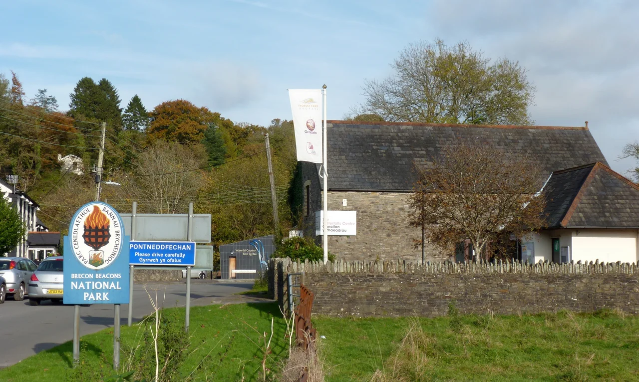 The Waterfalls Centre, main building centre right, provides information on the waterfalls of the River Neath tributaries, Brecon Beacons National Park and the Fforest Fawr Geopark. In the foreground is the boundary marker for the National Park and the boundary between Powys and Neath Port Talbot. On the extreme left is the historic Angel Inn, Pontneddfechan.