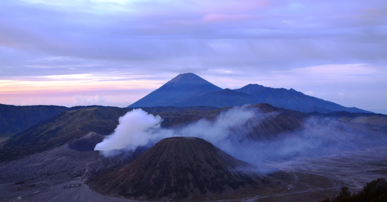 Gunung Batok, viewed from the seas of sand on the caldera of the Bromo complex in East Java, Indonesia. Grasses and shrubs can be seen growing on the few soil patches among the sands, while the mountain is littered with prominent trees.