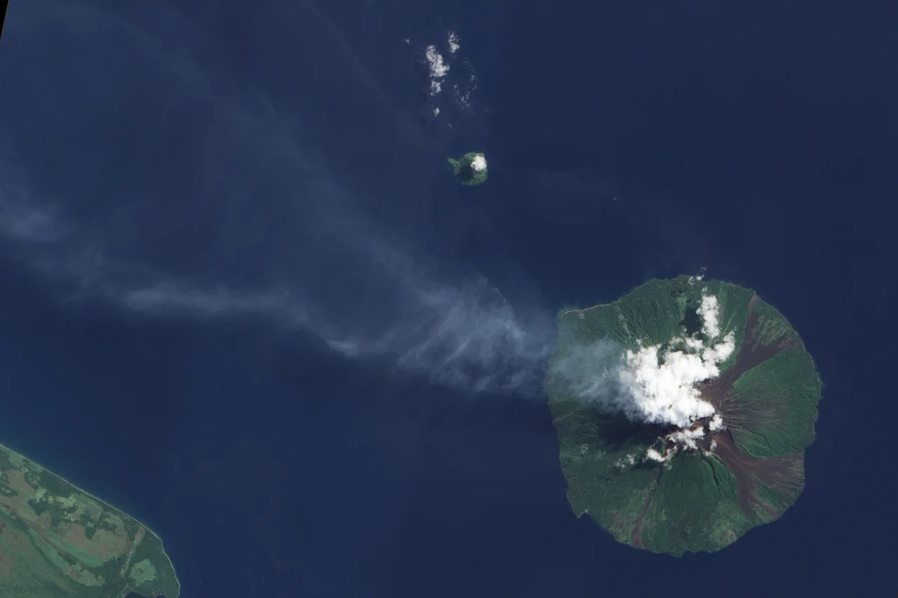 Bright white clouds hover over the volcano’s summit. Clouds often collect over peaks, but these clouds could result from water vapour released by the volcano. Slightly darker in colour, a pale blue-grey plume blows west-north-west from the summit and over the Bismarck Sea. Just 10 kilometres across, Manam is a stratovolcano composed of alternating layers of ash, lava, and rocks from prior eruptions. The circular island sports four radial valleys spaced roughly 90 degrees apart, and these valleys have historically channelled lava and pyroclastic flows—composed of hot rock, gas and ash—sometimes all the way to the coast. Evidence of earlier volcanic activity appears as rivulets of rock that interrupt the vegetation coating much of the island. Although clouds hide the summit in this image, the summit is known to support two craters devoid of vegetation.