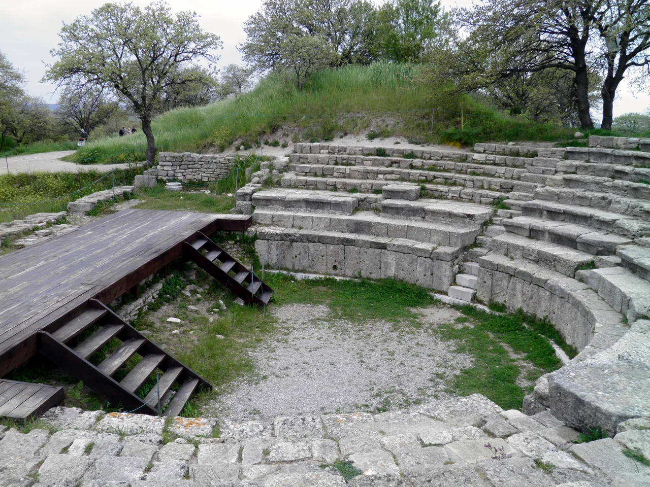 Hadrianic Odeon in Troy IX, small Theatre renovated by Hadrian in 124 AD.
In the early second century AD the provincial city of Ilion built a new theater unlike any architecture it had previously realized. At that time Roman Ilion was still best known for the large Hellenistic sanctuary of Athena (begun ca. 230 BC) with its imposing white marble temple complex bedecking the city’s acropolis. In contrast, the new theater was very small (perhaps the smallest in the whole Aegean area) and had an opulent scenae frons, or stage building. It was first discovered by Heinrich Schliemann’s architect and successor, Wilhelm Dörpfeld, in 1893, but received scant attention from archaeologists at Troy until recently (Aylward 2000, p. 138). Because of its diminutive size the current archaeologists from the University of Cincinnati name it an “odeion,” place for recitation of odes.
Briefly described, the Odeion of Ilion had a two-storey scenae of very limited depth, even including the aediculated bays. The foundation tells us how limited in depth the whole construction was, and it also shows an irregular column spacing mirrored about the center. The center opening is slightly wider than the pairs of openings to either side, and the pairs of columns which frame each opening are spaced even closer together. This inflected spacing of the vertical elements tells us the projecting and receding sections of entablature repeat in the upper storey in alignment (or stacking) with the lower entablature. This is in noticeable contrast to the trend since the late first century BC in Miletus and Ephesos where the spacing of the vertical elements is uninflected, thus allowing the projecting entablatures to alternate (or offset) from storey to storey without gross distortion of proportions. A secondary effect of the “in alignment” type of scheme is a greater inflection toward and emphasis of the center; at Ilion the Odeion has a large broken pediment spanning the center three bays at the upper storey, and at the lower storey entablature level there is no concavity, as expected by the normal rules. The upper storey also confounds the rules by including three different orders: Ionic for the major columns, Pergamene for the center bay, and Corinthian for the secondary niches. The whole scheme was a carefully orchestrated composition in white marble elements juxtaposed to colored marbles (blue-grey, violet, pink, coral, red, beige, green and yellow).

The emperor Hadrian visited the city in 124. He appears to have ordered new repairs and may have redecorated the odeon, where his statue has been found. His visit marks the beginning of a golden age, with the construction of Roman baths, a fountain (nymphaeum), and an aqueduct. Substantial parts of it survive and can be seen near Kemerdere.