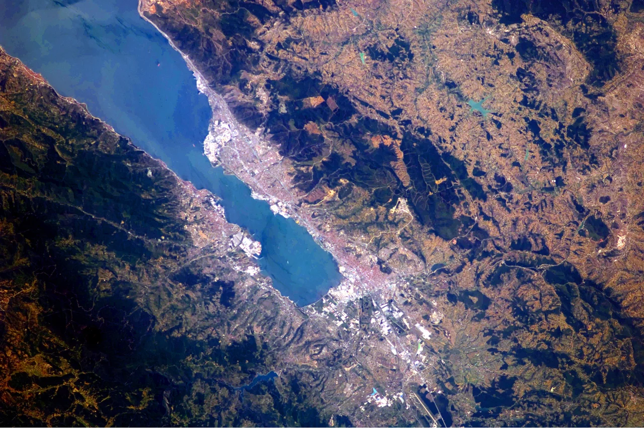 This astronaut photograph highlights the metropolitan area of Izmit along the northern and eastern shores of the Sea of Marmara, at the end of the Gulf of Izmit. Commercial and industrial centres—including petroleum refineries and automobile factories—are recognizable by large structures with white rooftops. The smaller city of Gölcük on the southern shoreline of the Gulf is the location of a Turkish naval facility and another automobile factory. Both urban areas are built primarily on flat lowlands adjacent to the Gulf, with green vegetation marking highland areas to the north of Izmit and south of Gölcük.