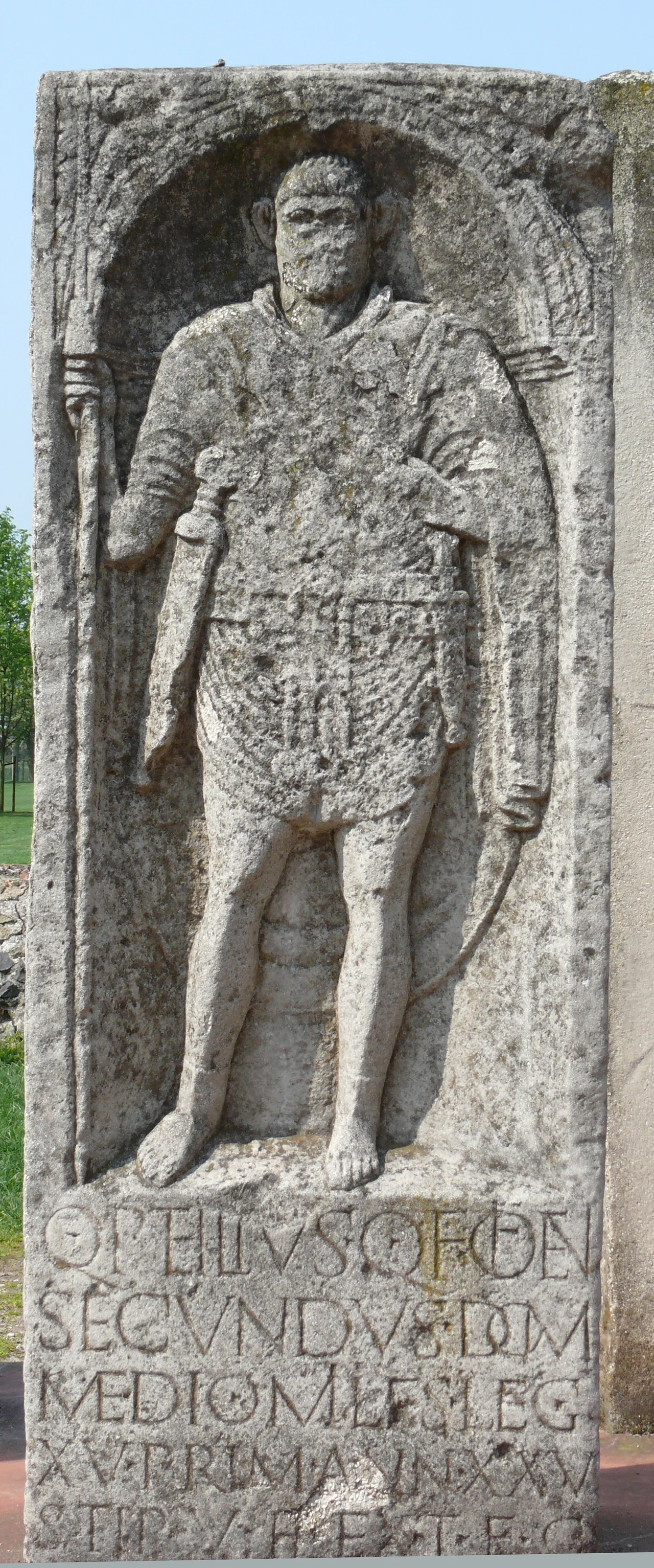 Photo taken in APX, the archeological park in Xanten. For the Latin text, see German version.
Quintus Petilius Secundus, son of Quintus from the tribe of the Oufentina from Milan, soldier of the 15th Legion Primigenia, 25 years old, 5 years in service, the heir has erected this gravestone in compliance with the testament.
The 15th Legion was stationed along with the 5th, in Xanten since 13 AD. Both legions built their camp Vetera in stone. The camp was destroyed by the Bataves during the 69/70 revolt.

Petilius is wearing a tunica and a shoulder cape. He is armed with a lance, a sword and a dagger.