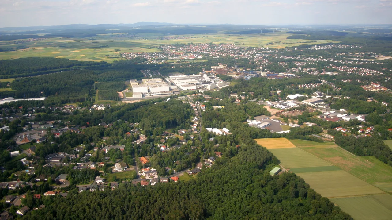 Stadtallendorf im Jahr 2004, fotografiert aus Richtung Westen. Im Foto zu sehen: Das Industriegebiet DAG mit dem Ferrero-Werk in der Bildmitte, der alte Ortskern ganz hinten, die Herrenwaldkaserne am rechten oberen Bildrand. Auf dem Foto wird die von großen Waldflächen durchzogene Bebauung deutlich.