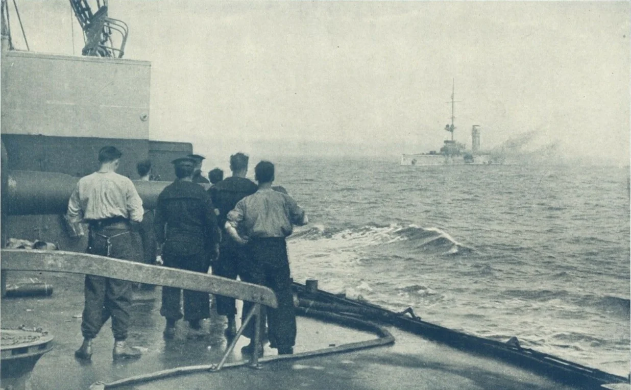 Titleː A Remarkable Camera Record of the Sinking of the German Cruiser Mainz off Heligoland August 28th 1914. A photograph taken from the deck of a British cruiser just before the Mainz sank, In the foreground British sailors watch the Mainz on fire.
The text indicates the photo was taken from the deck of one of the British cruisers present, and shows a British gun crew standing by their 6 inch gun. The Mainz is seen on fire, minus two after funnels and a mast, shortly before heeling over and sinking.
