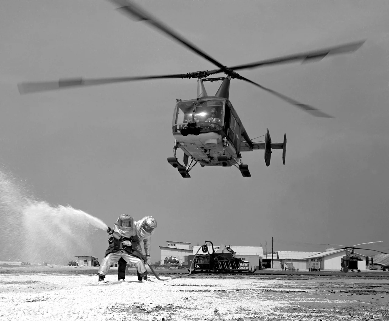 U.S. Air Force fire protection specialists fight a fire as an HH-43B Huskie helicopter hovers overhead to create a continuous downward air current, assisting the crew members. The crew is assigned to Detachment 8, 38th Aerospace Rescue and Recovery Squadron, Cam Ranh Bay Air Base, Vietnam.